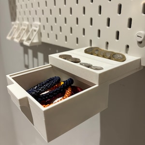 May include: A white plastic pegboard accessory featuring a small pull-out drawer and a top tray. The drawer contains several braided cord bracelets in navy blue and orange. The top tray holds a collection of various metallic coins.