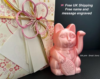 May include: A silk pink Maneki Neko cat figurine, approximately 3.5 inches tall, with a raised paw and holding a coin. The figurine is next to a decorative floral gift wrap.