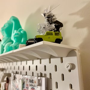 May include: A white shelf displays a collection of items. A lime green toy SUV sits next to a clear robot figure. A turquoise sculpture and other small items are also on the shelf. The shelf is mounted on a white pegboard.