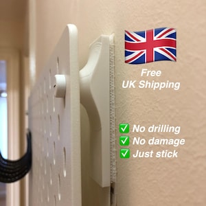 May include: A white pegboard system mounted on a beige wall. The system includes a mounting bracket and a pegboard with evenly spaced holes. Text on the wall reads "No drilling, No damage, Just stick."