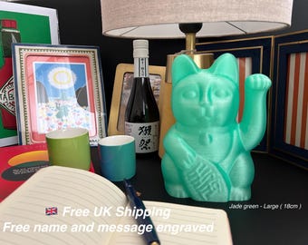 May include: A jade green, 18 cm tall, Maneki Neko cat figurine with a raised paw, holding a fish. The figurine is next to a bottle of sake, two ceramic cups, and a framed picture. A notebook and pen are in the foreground.