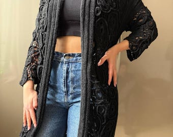 May include: A black, open-front cardigan with intricate crochet detailing. The long-sleeved garment features a flowing design with a textured pattern. The cardigan is worn over a black top and blue jeans.