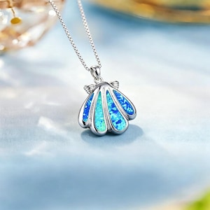 May include: A silver necklace with a seashell pendant. The pendant is a shell shape with a blue and turquoise opal-like stone. The chain is a delicate silver chain. The background is a soft blue.