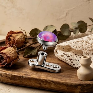 May include: A disassembled dermal anchor jewelry piece. The top features a round, iridescent purple and pink stone set in a silver bezel. The base is a silver, flat plate with three holes and a central post. The jewelry is designed for body piercing.