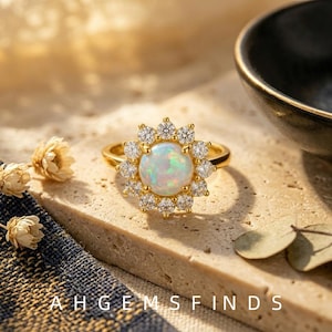 May include: A gold ring featuring a central round iridescent opal cabochon surrounded by a halo of twelve small, clear, round-cut stones. The ring band is thin and polished gold. The text AHGEMSFINDS is printed at the bottom of the image.