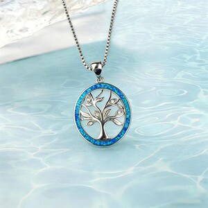 May include: Silver-toned necklace featuring a circular pendant with a tree of life design. The tree is detailed with leaves and small crystals, set against a blue opal-like background. The pendant hangs from a delicate chain.