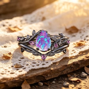 Puede incluir: Un juego de anillos negros con una gran piedra de ópalo ovalada e iridiscente en tonos morados y azules. El anillo está adornado con piedras rosas más pequeñas y un diseño de hojas. El conjunto está sobre una superficie de madera.