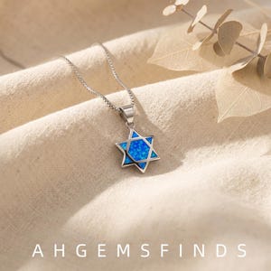 May include: A silver-toned Star of David pendant necklace featuring a vibrant blue opal inlay. The six-pointed star charm is suspended from a delicate box chain. The text AHGEMSFINDS is printed in white at the bottom of the image.