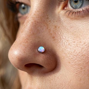 May include: Close-up of a nose with a small, round opal nose stud. The stud is set in a silver bezel and has a pearlescent, multi-colored appearance. The skin around the nose has freckles and is slightly out of focus.