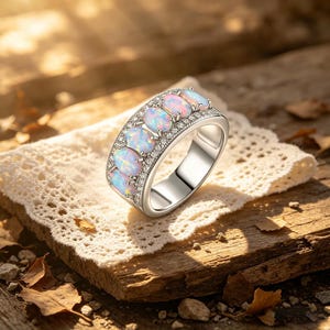 May include: A silver ring with five oval opals, displaying a play of colours. The opals are set in a row, with smaller clear gemstones on either side. The ring is displayed on lace and a wooden surface.