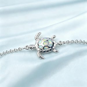 Op de afbeelding: Een zilverkleurige armband met een schildpadhanger. De schildpad heeft een opaalachtig midden en een gedetailleerd ontwerp van vinnen en kop. De armbandketting is delicaat. De achtergrond is zachtblauw.