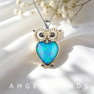 Könnte beinhalten: Eine silberfarbene Eulen-Halskette mit einem herzförmigen blauen Opal-Körper, zwei schwarzen runden Augen und goldfarbenen Akzenten um die Augen, die mit kleinen klaren Steinen besetzt sind. Der Anhänger hängt an einer zierlichen silberfarbenen Kette.