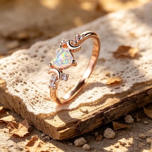 May include: Rose gold ring featuring a heart-shaped opal gemstone, accented with small clear crystals and crescent moon details. The ring is set on a wooden surface with a lace doily.