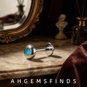 Könnte beinhalten: Ein silberfarbenes Barbell-Piercing mit einem glatten, polierten Metallschaft und zwei abgerundeten Enden, die jeweils mit einem leuchtend türkisfarbenen, kreisförmigen Stein besetzt sind. Der Text AHGEMSFINDS steht unten im Bild.