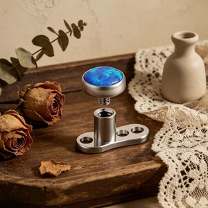 May include: A silver-toned dermal anchor with a blue and silver gemstone top. The base has a flat, rectangular shape with three holes. The gemstone has a blue and white marbled pattern. The anchor is designed for body piercing.