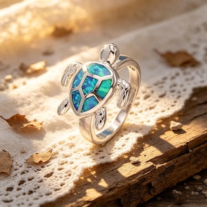 May include: A silver-colored ring featuring a turtle design. The turtle's shell is inlaid with a vibrant blue and green gemstone. The ring is set against a rustic wooden surface and a white lace cloth.