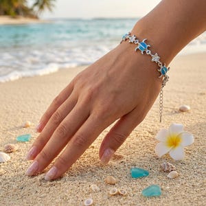 Op de afbeelding: Een zilveren armband met blauwe zeeschildpad- en sterbedels. De armband zit om een pols, met een strand en oceaan op de achtergrond. De armband is ongeveer 18 cm lang.