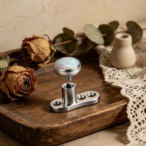 May include: A disassembled dermal anchor with a round, iridescent opal top and a silver-colored base. The base has a rectangular shape with three holes. The opal is set in a silver bezel. The anchor is designed for body piercing.