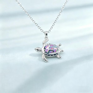 May include: A silver-colored necklace featuring a turtle pendant. The turtle pendant has an iridescent, multi-colored opal-like center and detailed silver accents. The necklace chain is delicate and hangs against a light blue background.