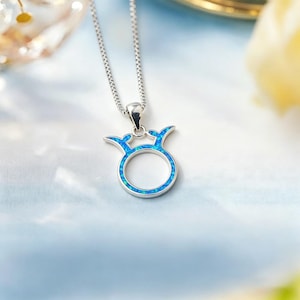 May include: A silver-colored pendant necklace featuring a Taurus zodiac symbol. The pendant has a blue opal-like center and is suspended from a delicate chain. The design is a stylized representation of the Taurus symbol.