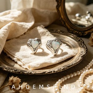 Puede incluir: Dos anillos de tono plateado con una piedra iridiscente en forma de corazón engastada en un marco de metal texturizado y orgánico. Los anillos se muestran sobre una tela, con el texto AHGEMSFINDS visible en la parte inferior.