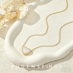 Op de afbeelding: Een gouden ketting met een hartvormige hanger met een gladde witte steen in het midden, omringd door een rand van kleine heldere steentjes. De ketting ligt op een wit gebogen oppervlak met de tekst AHGEMSFINDS onderaan in de afbeelding.