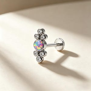 May include: A silver-colored lip piercing with a cluster of clear, round gemstones and a central, iridescent opal-like stone. The design features a flat back and a threaded post for secure placement. The jewelry is designed for body modification.
