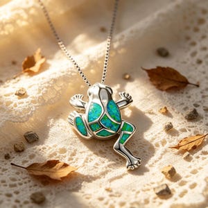 May include: A silver frog pendant necklace with a turquoise and green opal inlay. The frog is detailed with outstretched arms and legs, and is suspended from a delicate chain. The necklace is displayed on a white lace surface with scattered leaves and small stones.