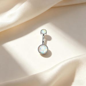 May include: A silver belly button ring with two opal-like, round stones. The larger stone is at the bottom, and the smaller stone is at the top. The stones have a white and iridescent appearance. The ring is displayed on a white, satin-like fabric.