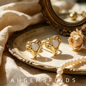 Puede incluir: Un par de pendientes dorados con un marco de metal orgánico y texturizado que rodea una piedra de ópalo iridiscente en forma de corazón. El texto AHGEMSFINDS es visible en la parte inferior de la imagen.