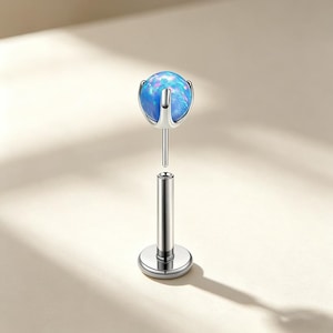 May include: A silver-colored lip piercing stud with a blue opal-like ball. The ball is held in place by a three-pronged setting. The post is cylindrical with a flat, round base.