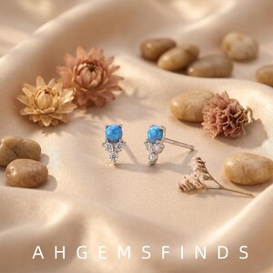 May include: A pair of silver stud earrings featuring a round blue opal cabochon at the top, accented by three small clear round-cut stones arranged in a triangular cluster below. The text AH GEMS FINDS is printed at the bottom of the image.