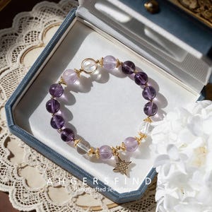 May include: A handcrafted bracelet featuring amethyst and clear crystal beads, accented with gold-colored metal details. The bracelet includes a star charm and is presented in a blue jewelry box. The text "AHGERSFINDS Handcrafted Jewellery" is visible.