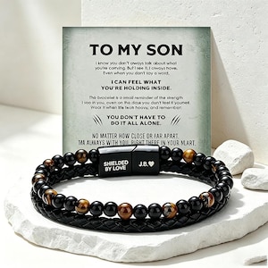 Peut inclure: Un bracelet noir composé de perles en œil de tigre et d'onyx, associé à un bracelet en cuir noir. Le bracelet est doté d'un fermoir magnétique avec le texte "SHIELDED BY LOVE" et les initiales "J.B.". Une carte avec le texte "TO MY SON" est en arrière-plan.