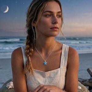 May include: A silver necklace with a small, blue turtle pendant. The pendant is centered on a delicate chain. The image is set against a beach backdrop with a crescent moon in the sky.