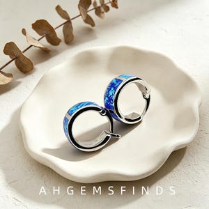 May include: A pair of silver hoop earrings featuring a vibrant blue inlaid stone pattern. The earrings have a polished metallic finish and a hinged closure mechanism. The text AHGEMSFINDS is printed at the bottom of the image frame.