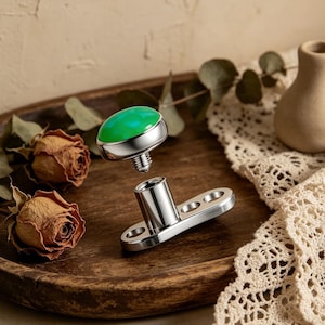 May include: A silver dermal anchor with a bright green, rounded gem. The anchor is disassembled, with the gem and threaded post separated from the base. The base has two holes for securing the anchor.