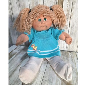 May include: A doll with light brown yarn hair styled in two pigtails, green eyes, and a tan complexion. The doll wears a bright turquoise knit dress featuring a small yellow duck graphic and white horizontal stripes, paired with plain white leggings.