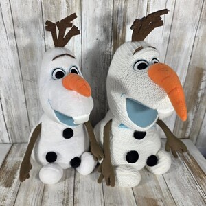 May include: Two plush snowman toys featuring white bodies, brown stick-like arms, brown twig hair, and orange carrot noses. The left snowman has a smooth white fabric body, while the right snowman has a textured, knit-style white fabric body.