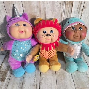 May include: Three Cabbage Patch Kids dolls. The left doll wears a purple unicorn costume with a silver horn and star pattern. The center doll wears a brown lion costume with a red mane. The right doll wears a light blue shark costume.