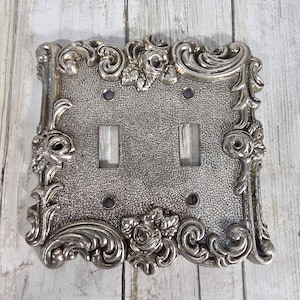 May include: A silver-toned double light switch plate featuring a textured, pebbled center surface and an ornate, raised border decorated with sculpted rose blossoms and swirling scrollwork patterns, including four screw holes for mounting.