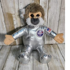 May include: A plush monkey toy with brown and white fur, wearing a metallic silver astronaut suit. The suit features a circular NASA logo patch on the chest and a small United States flag patch on the left sleeve. The monkey has large black eyes.