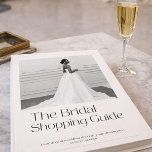 May include: A white book titled "The Bridal Shopping Guide" with a photo of a bride in a wedding dress. A glass of champagne sits next to the book on a marble surface. The book's subtitle reads "Your dream wedding dress at your dream pice".