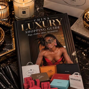 May include: A hardcover book titled China Luxury Shopping Guide by Shirley Bekker, featuring a cover image of a person in a red dress and black mask, surrounded by various miniature luxury brand shopping bags and boxes on a dark surface.