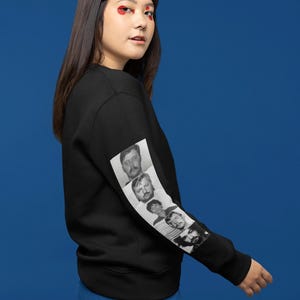 May include: Black sweatshirt featuring a series of black and white mugshot images on the left sleeve. The images are of men with facial hair. The sweatshirt is worn by a person with long dark hair and blue trousers.