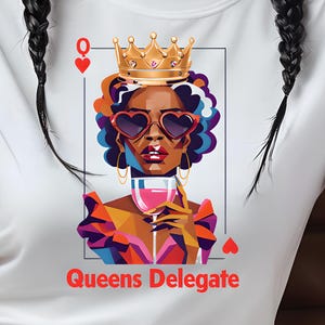 May include: White t-shirt featuring a graphic of a queen with a crown, heart-shaped sunglasses, and a glass of wine. The text "Queens Delegate" is printed below the image. The model is wearing white pants and holding a coffee cup.