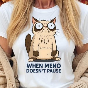 May include: White t-shirt featuring a cartoon cat with a grumpy expression. The cat is light brown with dark brown accents. The text "WHEN MENO DOESN'T PAUSE" is printed below the cat in dark blue. The background includes fall foliage and pumpkins.