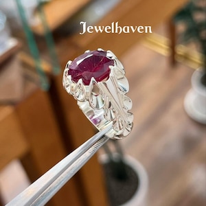 Könnte beinhalten: Ein silberner Ring mit einem großen, oval geschliffenen, tiefroten Edelstein. Der Ring hat ein verziertes, blumeninspiriertes Design um das Band und die Edelsteinfassung. Der Text "Jewelhaven" ist sichtbar.