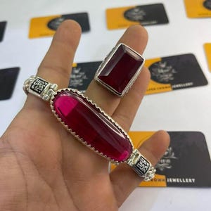 Può includere: Un set di bracciale e anello in argento con grandi pietre preziose rosso rubino rettangolari. Il bracciale ha una pietra ovale, mentre l'anello ha una pietra quadrata. Entrambi i pezzi presentano dettagli in argento.