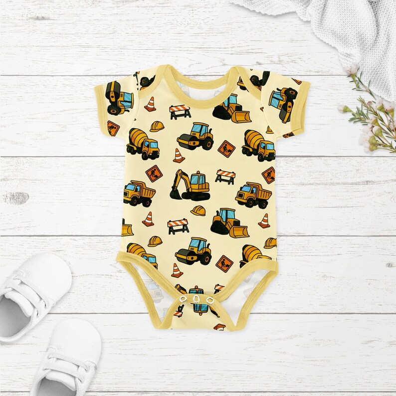 May include: A pale yellow baby bodysuit with short sleeves and a construction vehicle pattern. The pattern includes images of trucks, excavators, cement mixers, traffic cones, and hard hats. The bodysuit has yellow trim and poppers at the bottom.